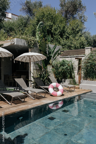 Boutique Hotel Pool Terrace with Sun Loungers and Balinese Umbrellas. Stylish boutique hotel pool area with sunbeds, tasseled Balinese umbrellas and a pink striped inflatable ring on a wooden deck. 