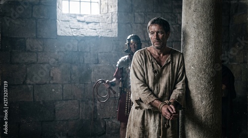 Apostle Paul leaning against stone pillar with hands bound in rope before Roman guard, Book of Acts New Testament biblical prison scene.