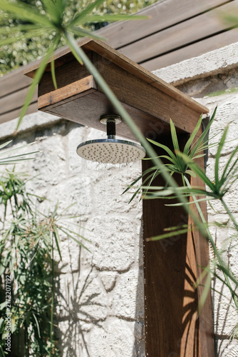 Outdoor Rain Shower with Running Water. Close-up of a poolside garden shower in action: sparkling water drops falling in sunlight, rustic wood, stone textures, lush tropical greenery of Mediterranean.