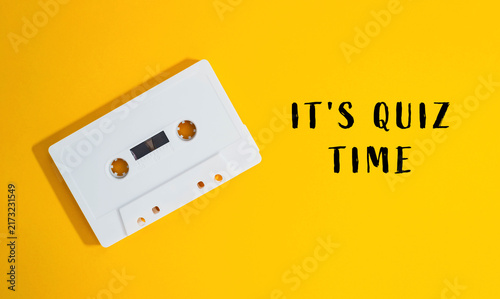 White cassette tape placed on a vibrant yellow background with the text 'It's Quiz Time' in bold letters, suggesting a retro themed quiz or trivia event.