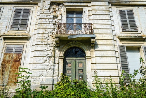 Photography Entrée et le premier étage d'une demeure bourgeoise en ruine, montrant une maçonnerie en pierre calcaire écroulée, un balcon ouvragé rouillé et une porte envahie par la végétation
