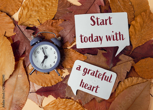 An alarm clock placed on a bed of autumn leaves alongside speech bubbles with the motivational quote 'Start today with a grateful heart', suggesting mindfulness and gratitude.