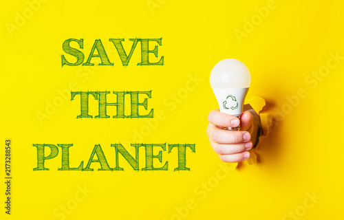 A hand holding an LED bulb with a recycling symbol, emerging from a yellow background, accompanied by the text 'Save the Planet', emphasizing eco friendly energy solutions.