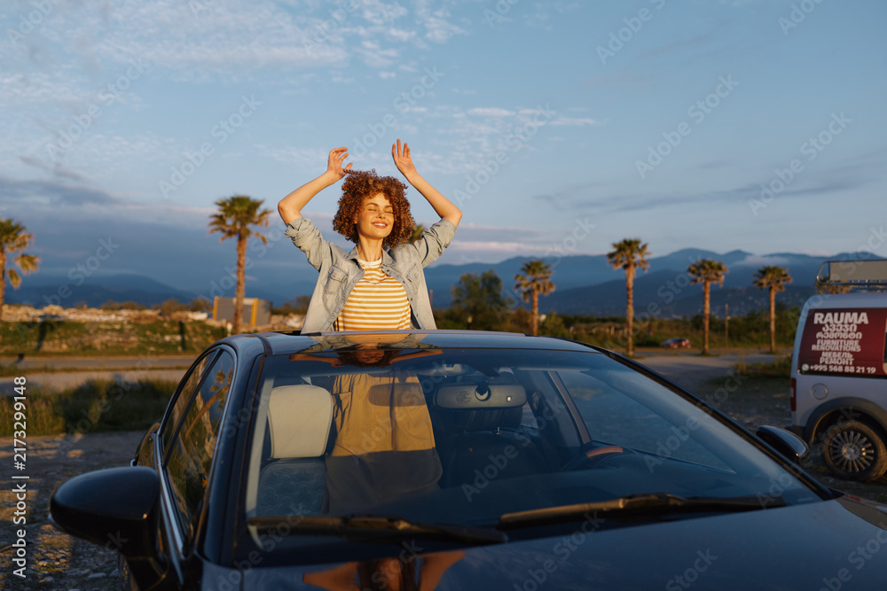 Fototapeta premium A young woman with curly hair stands through a sunroof, arms raised, in a bright desert scene with palm trees, conveying freedom and carefree mood.