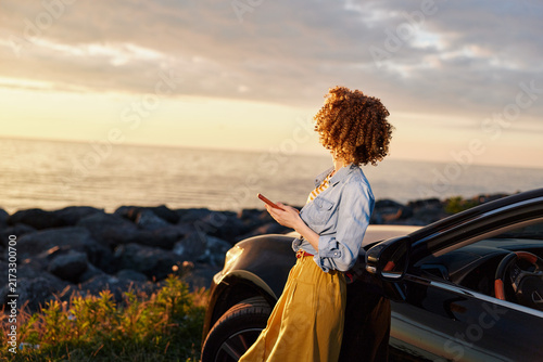 Fotografie A stylish young woman with curly hair stands beside a sleek car on a rocky coast at sunset, wearing a denim shirt and bright yellow skirt while glancing at her phone