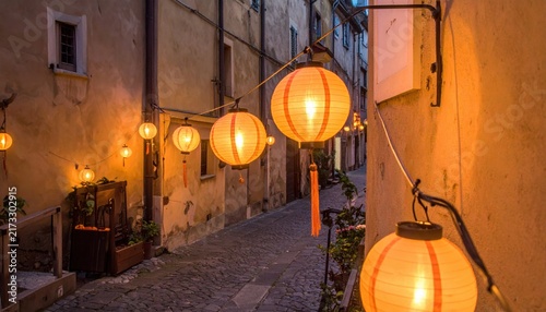 Warm orange lanterns glow on a cobblestone alleyway at night, illuminating a charming village street