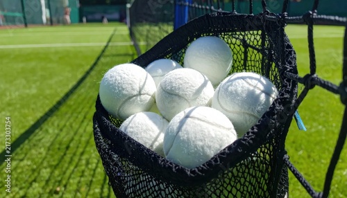 White Tennis Balls Overflowing From A Black Net Basket On A Lush Green Court