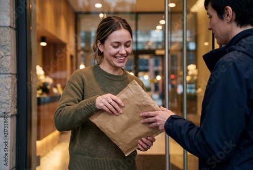 Friendly woman receiving a small cardboard delivery package from a courier at the entrance of a modern quiet apartment building doorway