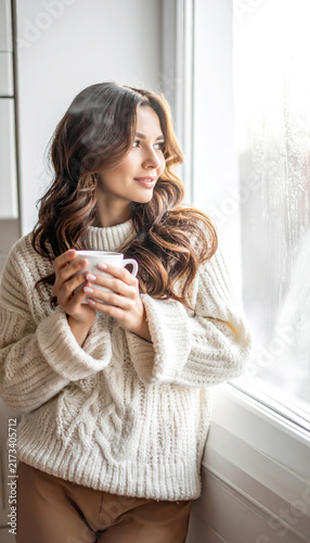 Junge Frau im Strickpullover genießt heißen dampfenden Kaffee am Fenster im Morgenlicht