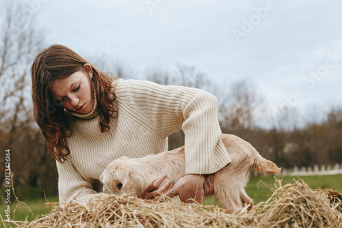 Foto A woman in a cream knit sweater bends beside hay, gently guiding a young goat kid in a sunny outdoor farm scene
