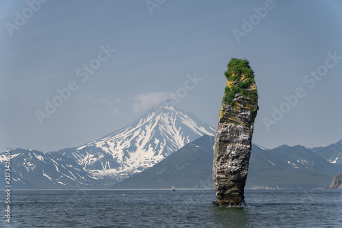 Bild auf Leinwand Tall sea stack rises from Avacha Bay with snow-covered volcano and mountain range behind in Kamchatka, Russia
