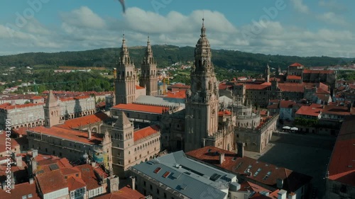 Majestic aerial view of the Santiago de Compostela Cathedral dominating the historic cityscape, with its intricate Baroque towers rising above terracotta rooftops under a bright, sunny sky, capturing