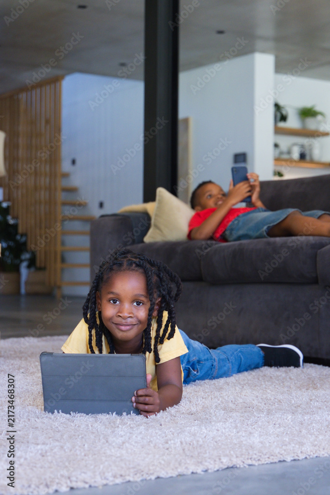 Naklejka premium African American children lying on plush rug in living room, holding tablet and smartphone