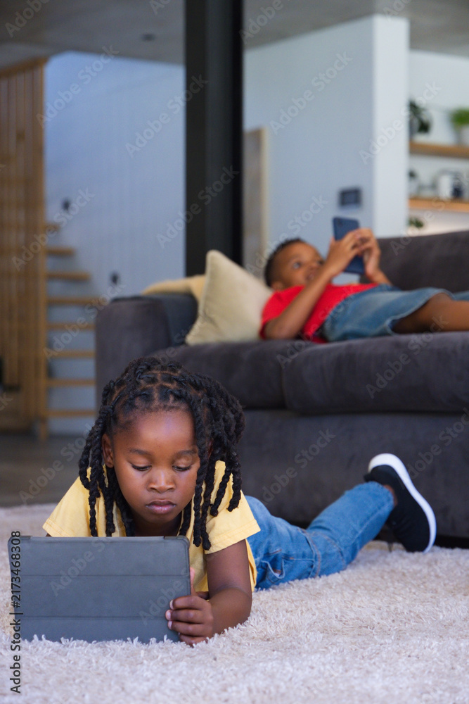 Naklejka premium Teenage girl lying on rug using tablet, sibling reclining on sofa with smartphone in living room