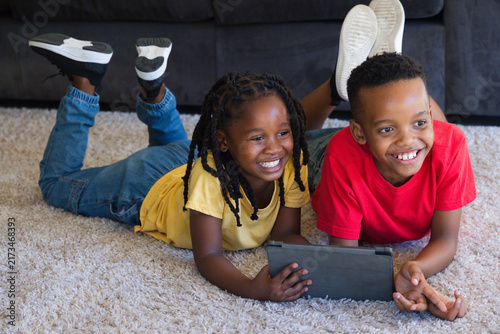 African American youth siblings lying on plush carpet at home sharing tablet and sneakers nearby