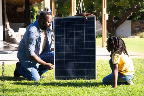 African American father and son kneeling and holding solar panel on backyard lawn near porch