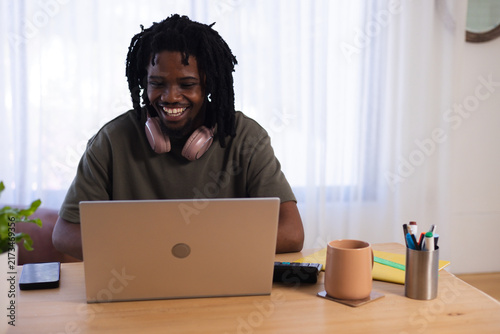 Adult man sitting at wooden table working on laptop wearing pink headphones, phone and mug visible