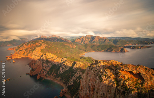 Dramatic sunset over the rugged coast of Corsica, France, with rocky mountains, Mediterranean Sea and sweeping clouds captured in warm golden light and long exposure.