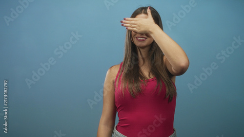 Bild auf Leinwand Young woman covers her eyes with hand while smiling in studio wearing red tanktop and jeans; playful shy moment