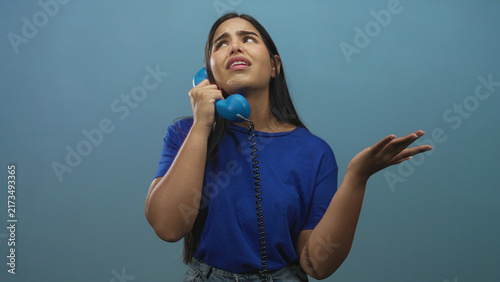 Fotografie Young hispanic woman holding blue corded telephone to her ear with hand raised in pleading gesture in studio; despair longing
