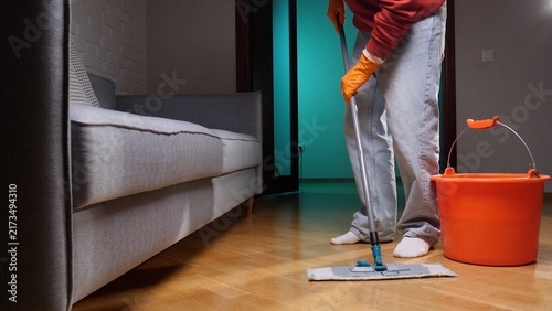 Obraz na plátne Person in orange gloves mopping a wooden living room floor in low evening light,