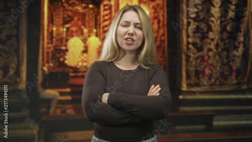 Bild auf Leinwand Blonde woman with folded arms and a grimace, standing before an ornate altar with robed priests in a church interior; disgust defiance