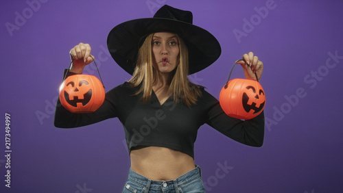 Photography Woman in witch hat holds two jack o lantern buckets by handles, puckered lips and exposed midriff against purple studio wall; playful mischief