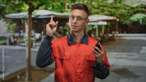 Konstfotografi Young blond man in orange coverall holding a walkietalkie and points finger up on a street cafe terrace while wearing glasses and looking focused; alert readiness