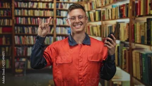 Konstfotografi Man mechanic in orange coverall holding a walkie talkie and making rock horns gesture with his hand in a building library; playful joy