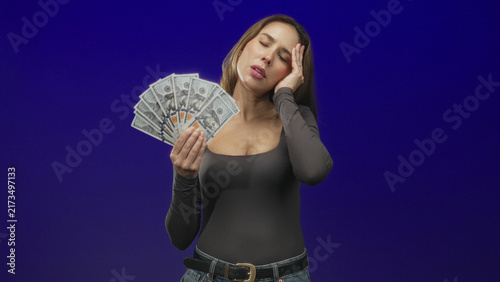 Cuadro en lienzo Woman holding a fan of hundred dollar bills with hand to temple, closed eyes and pained expression, wearing fitted top and jeans in studio; financial stress