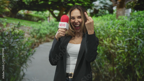 Hispanic woman reporter in blazer holding red news microphone makes explicit middle finger gesture while smiling on a leafy street path; defiance.