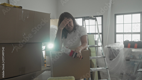 Foto Woman leaning on cardboard moving box holding tape dispenser with hand on forehead for headache beside ladder and covered furniture in building; moving stress