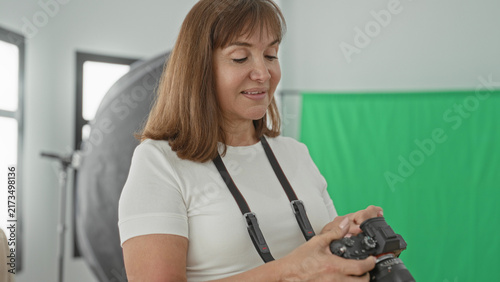 Фотография Woman adjusting camera with hands and strap, green screen backdrop visible in studio; calm professionalism