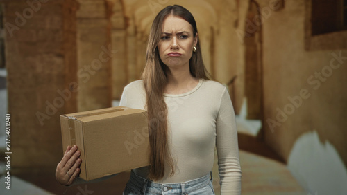 фотографія Young hispanic woman holding delivery box with pouting expression in historic building corridor; quiet frustration