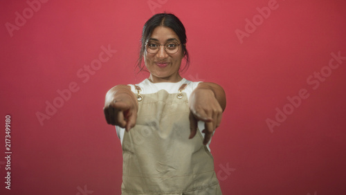 Cuadro en lienzo Woman wearing apron smiles and points finger to camera against red studio backdrop; cheerful playful