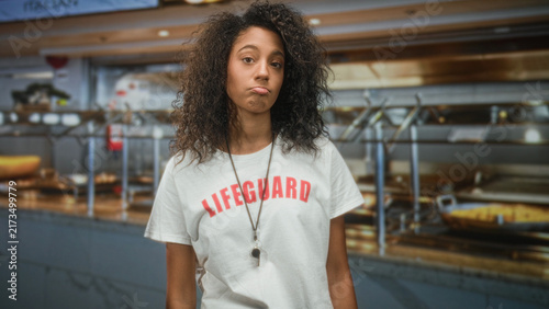 Принти на полотні Teen woman in lifeguard tshirt pouting lips with whistle necklace at cafeteria serving line inside building; boredom duty