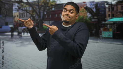 Canvas Print Hispanic man pointing index fingers and shrugging shoulders on an urban street near a food kiosk, wearing knit sweater and smirk; uncertainty streetlife