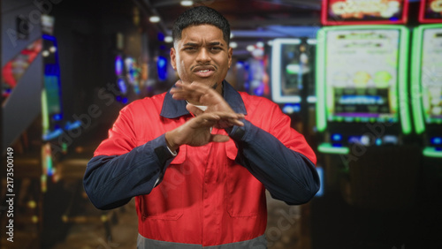 Canvas Print Man in worker uniform and red jacket pinches nose with hands, grimacing and covering face, beard visible, standing by slot machines in casino; smell disgust