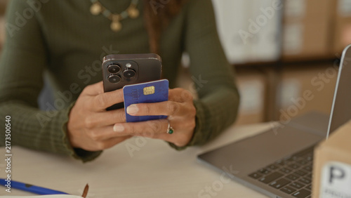 Quadro su tela Woman holding smartphone and creditcard in hands at a studio desk with parcels, laptop and notebook, tapping to process orders; small business focused