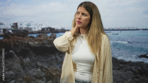 Obraz na płótnie Woman touching her neck with one hand, wincing from neck pain while posed against a rocky seaside backdrop, photographed in studio; travel discomfort