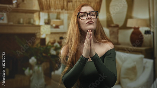 Konstfotografi Redhead woman wearing black framed glasses presses bare hands together in studio living room set with lamp, vase, couch and fireplace visible; pleading