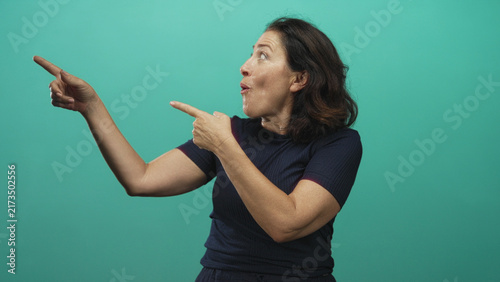 Quadro su tela Woman pointing index fingers with surprised open mouth and animated expression in studio teal backdrop; playful surprise