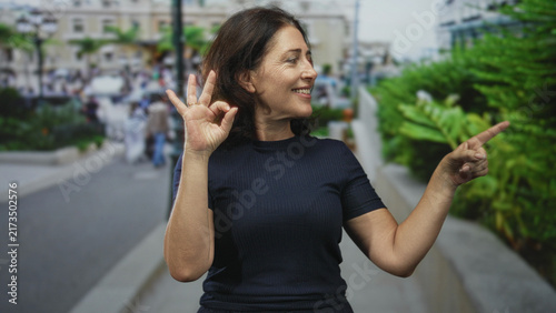 Fotografía Middle age woman makes ok sign with left hand and points right finger on busy urban street; confidence approval gesture