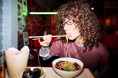 Young adult Asian woman eating ramen noodles with chopsticks in Asian restaurant. Image represents food culture and is suitable for culinary blogs and restaurant marketing, close up shot