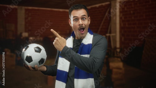 Foto Hispanic man with moustache holding a soccer ball and pointing finger in a building; excitement passion celebration