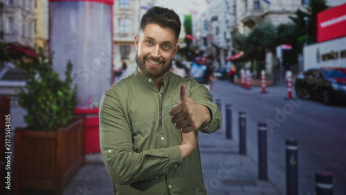 Man with beard in green shirt smiling with arms crossed and occasional thumbs up gesture, bare right hand visible, standing on city street with bollards and planter; cheerful confidence.