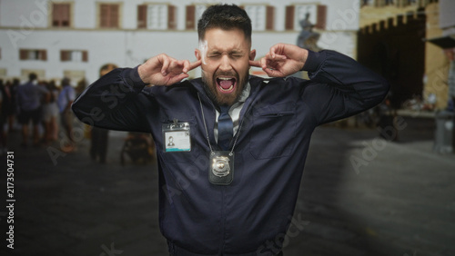 фотографія Man police detective covering ears and shouting with badge and id visible on street during crowd scene; frustration overload