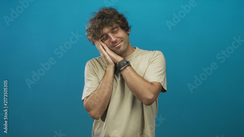 Photography Young hispanic man in beige t shirt presses both hands to cheek with closed eyes and gentle smile against studio blue backdrop; sleepiness contentment