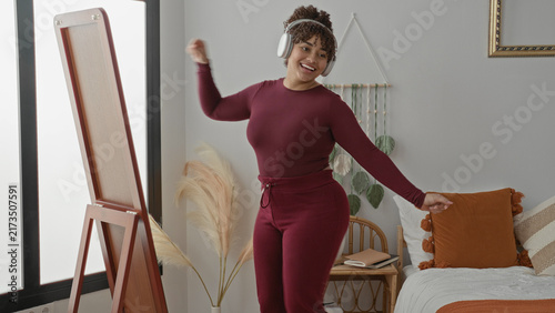 Obraz na plátne Woman dancing with headphones, smiling and arms raised beside a standing mirror and bed in a bright studio bedroom; joyful relaxation