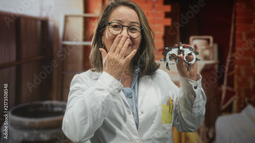 Bild auf Leinwand Hispanic senior woman with grey hair in white coat holds optometry trial frame while covering mouth with hand, posing in studio among props; playful curiosity